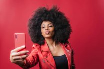 Femme afro-américaine souriante avec coiffure afro prenant autoportrait sur téléphone portable sur fond rouge en studio — Photo de stock