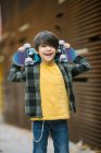 Menino alegre em roupas casuais sorrindo e olhando para a câmera enquanto estava de pé com skate atrás da cabeça na rua contra fundo borrado — Fotografia de Stock