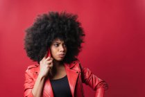 Excited African American female with Afro hairstyle and pouting lips browsing mobile phone on red background in studio — Stock Photo