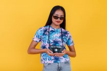 Happy Asian female in t shirt with tropical leaf print holding photo camera on yellow background in studio — Stock Photo