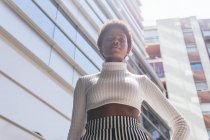 Снизу стильная самоуверенная афроамериканка держит за талию и смотрит в камеру, стоя рядом с современными многоквартирными домами в солнечный день в городе — стоковое фото