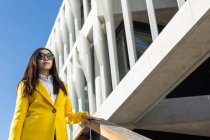 Mulher de negócios asiática com casaco amarelo e telefone inteligente andando na rua com edifício no fundo — Fotografia de Stock