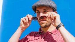 Young bearded guy wearing casual red polo shirt and cap looking away over sunglasses while standing against blue wall on street in summer day — Stock Photo