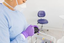 Doctora adulta en máscara uniforme y respiratoria con placa pequeña y pistola de agua durante el proceso de limpieza en la clínica - foto de stock