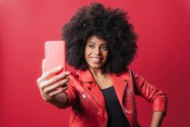 Smiling African American female with Afro hairstyle taking self portrait on mobile phone on red background in studio — Stock Photo