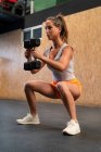 Все тело сфокусированной худенькой спортсменки делает приседания с тяжелыми гантелями во время тренировки в тренажерном зале — стоковое фото