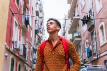 Низкий угол уверенного в себе молодого этнического туриста с рюкзаком и наушниками TWS, смотрящего в сторону, исследуя старые узкие улочки Барселоны — стоковое фото