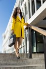Asiatica donna d'affari con cappotto giallo e smart phone a piedi per strada con edificio sullo sfondo — Foto stock