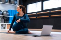 Corpo cheio de flexível jovem instrutor de ioga do sexo feminino fazendo metade senhor dos peixes posar na frente da tela do laptop durante a aula on-line no estúdio de fitness — Fotografia de Stock