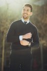 Imprenditore maschio barbuto sorridente in orologio da polso con taglio di capelli moderno guardando altrove in città in retroilluminazione — Foto stock
