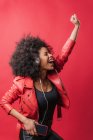 Entzückte Afroamerikanerin hört Musik über Kopfhörer und benutzt Mobiltelefon, während sie im Studio auf rotem Hintergrund tanzt — Stockfoto