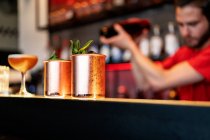 Cóctel de mula de Moscú en tazas de cobre servidas en el mostrador en el bar en el fondo de barman borroso con agitador - foto de stock
