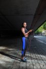 Vista laterale della bella donna afroamericana in abbigliamento sportivo che tiene la corda di salto e guarda la fotocamera mentre si trova sul marciapiede sulla strada della città — Foto stock