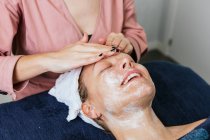 Врожай невизначений косметолог, який застосовує засоби для чищення обличчя на обличчі жіночого клієнта під час догляду за шкірою в салоні краси — стокове фото