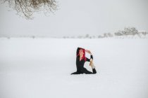 Спокойная женщина с розовыми волосами сидит в Eka Pada Raja Kapotasana на снежном зимнем поле и практикует йогу — стоковое фото