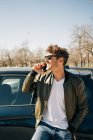 Happy male in sunglasses talking on cellphone while standing near automobile on sunny day — Stock Photo