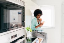 Moderna bella donna afroamericana con smartphone in mano seduto sul bancone della cucina guardando lontano a casa e mangiando mela — Foto stock