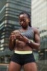Mujer afroamericana en ropa deportiva que navega por un teléfono inteligente moderno mientras está de pie sobre un fondo borroso de la calle de la ciudad durante el entrenamiento al aire libre - foto de stock