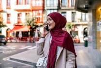 Mujer musulmana alegre en hijab y con bolsas de compras caminando por la calle y hablando en un teléfono inteligente - foto de stock
