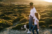 Full body side view of happy multiethnic woman and little girl with Border Collie dog walking together on trail among grassy hills in sunny spring evening — Stock Photo