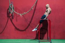 Вид збоку бородатий спортсмен робить вправи на брусах проти червоної стіни під час тренувань у спортзалі — стокове фото