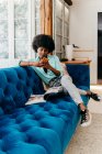 Молодая афроамериканка в повседневной одежде просматривает смартфон и читает журнал, отдыхая дома на удобном синем диване — стоковое фото