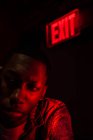 Молодой афроамериканец смотрит в сторону подсвеченной пластины Выход над головой в красном темном свете — стоковое фото