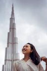 Bajo ángulo de sonriente joven hembra asiática en traje casual mirando hacia otro lado mientras que de pie contra la moderna torre de Burj Khalifa en Dubai en día nublado - foto de stock