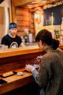 Seitenansicht einer zufriedenen Asiatin im Pullover, die lächelt, während sie einem Arbeiter, der an der Holztheke in der Ramen-Bar sitzt, den Löffel wegnimmt — Stockfoto