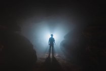 Silhouette di esploratore maschio anonimo in piedi da solo in buia stretta grotta rocciosa contro la luce incandescente dall'ingresso — Foto stock