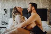 Jovem alegre e mulher sorrindo e abraçando enquanto sentados em uma cama confortável em casa juntos — Fotografia de Stock