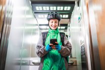 Low angle of glad female courier in uniform and helmet smiling and browsing cellphone in elevator while making delivery — Stock Photo