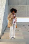 Low angle of ethnic male in vintage coat with Afro hairstyle text messaging on cellphone on urban staircase — Stock Photo