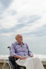 Macho sênior em cadeira de rodas no calçadão olhando para longe em contemplação e desfrutando de vista para o mar — Fotografia de Stock
