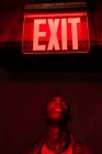 From below crop African American young male looking up on illuminated tablet Exit above head in red dark light — Stock Photo