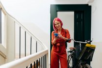 Jovem mulher elegante em terno vermelho com mochila usando smartphone enquanto está de pé com bicicleta na escada — Fotografia de Stock