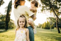 Nettes Vorschulmädchen lächelt und schaut in die Kamera, während es Mutter mit kleiner Schwester an den Händen während des Sommertages im grünen Park schmust — Stockfoto