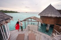 Von oben Rückansicht einer nicht wiedererkennbaren Frau im roten Kleid mit erhobenem Arm, die am Meer tanzt, mit einem gesichtslosen Partner in Freizeitkleidung, der es sich auf den Malediven gemütlich macht — Stockfoto