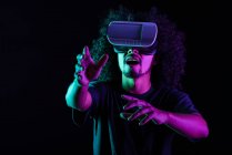 Homem latino animado com penteado afro e em óculos VR experimentando realidade virtual em fundo preto em estúdio com luzes de néon — Fotografia de Stock