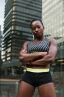Mujer negra positiva en ropa deportiva manteniendo los brazos cruzados y mirando a la cámara mientras está de pie sobre un fondo borroso de la calle de la ciudad - foto de stock