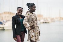 Задоволені стильними афроамериканськими жінками, які тримаються поруч і дивляться на камеру з задумливою посмішкою в парку в яскравий день. — стокове фото