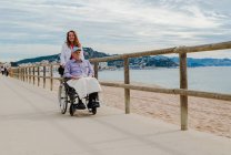 Encantada filha adulta empurrando cadeira de rodas com pai sênior e curtindo passeio ao longo do passeio perto do mar — Fotografia de Stock