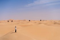 Giovane donna sorridente in abiti casual in piedi su dune sabbiose contro deserto durante il viaggio negli Emirati e agitando la mano — Foto stock
