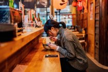 Side view of satisfied Asian woman in sweater smiling while taking spoon from worker sitting at wooden counter in ramen bar — Stock Photo