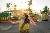 Contenuto femminile scattare autoritratto sul telefono cellulare divertendosi nel parco divertimenti in serata in estate — Foto stock