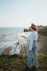 Вид на молодую женщину в стильной одежде и берете, стоящую на травянистом побережье возле песка и океана в солнечный день, рисуя картину кистью на холсте на мольберте — стоковое фото