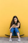 Молода жінка-кітсерфінг інтернет на мобільному телефоні, сидячи на скейтборді на прогулянці вдень — стокове фото