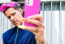 Hombre homosexual de moda con el pelo rosa y uñas de pechuga de colores tomando uno mismo tiro en el teléfono inteligente en la calle - foto de stock