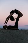 Vista lateral da silhueta da mulher flexível fazendo backbend e equilíbrio nas pernas do homem durante a sessão de acroyoga contra o céu do pôr do sol com a lua — Fotografia de Stock