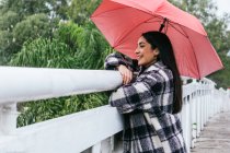 Side view of optimistic ethnic female with umbrella smiling and looking away while leaning on bridge railing on rainy day in park — Stock Photo